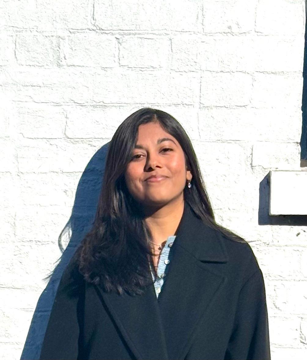 Anindita Shrivastava in a black coat against a white painted brick wall.